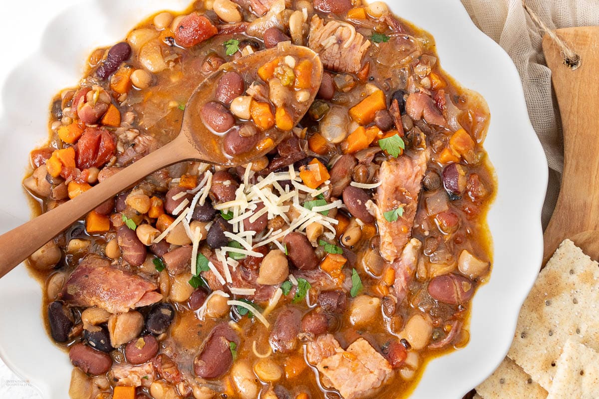A bowl of mixed bean and ham soup with chopped carrots, topped with grated cheese and parsley, a wooden spoon, and crackers on the side—one of the most comforting soups that showcases classic bean soup recipes.