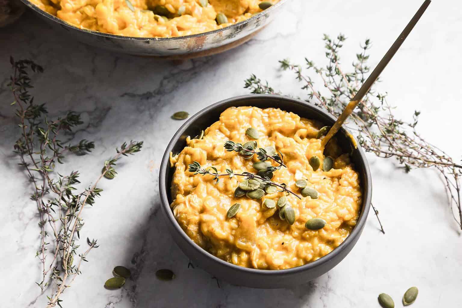 A bowl of creamy macaroni and cheese topped with pumpkin seeds and a sprig of thyme, with fresh thyme scattered on a marble surface nearby, makes for a cozy autumn dish perfect for anyone seeking new recipes or delicious pumpkin recipes.