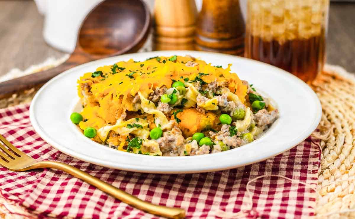 A plate of casserole topped with melted cheese, ground meat, peas, and noodles sits on a white plate, evoking the comforting flavors of classic grandma recipes. A fork and a glass of iced drink are placed nearby, completing this timeless autumn meal.