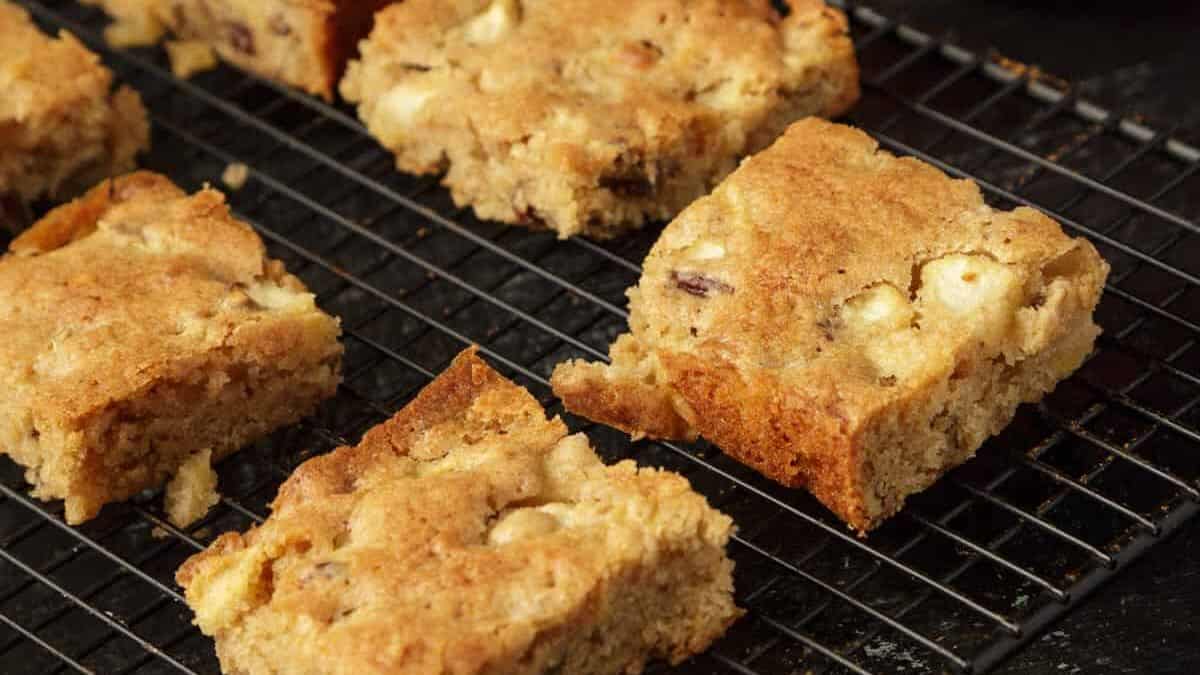 Five pieces of blondie bars with visible white chocolate chunks are cooling on a black wire rack—perfect as an easy dessert for kids or a cozy autumn treat.