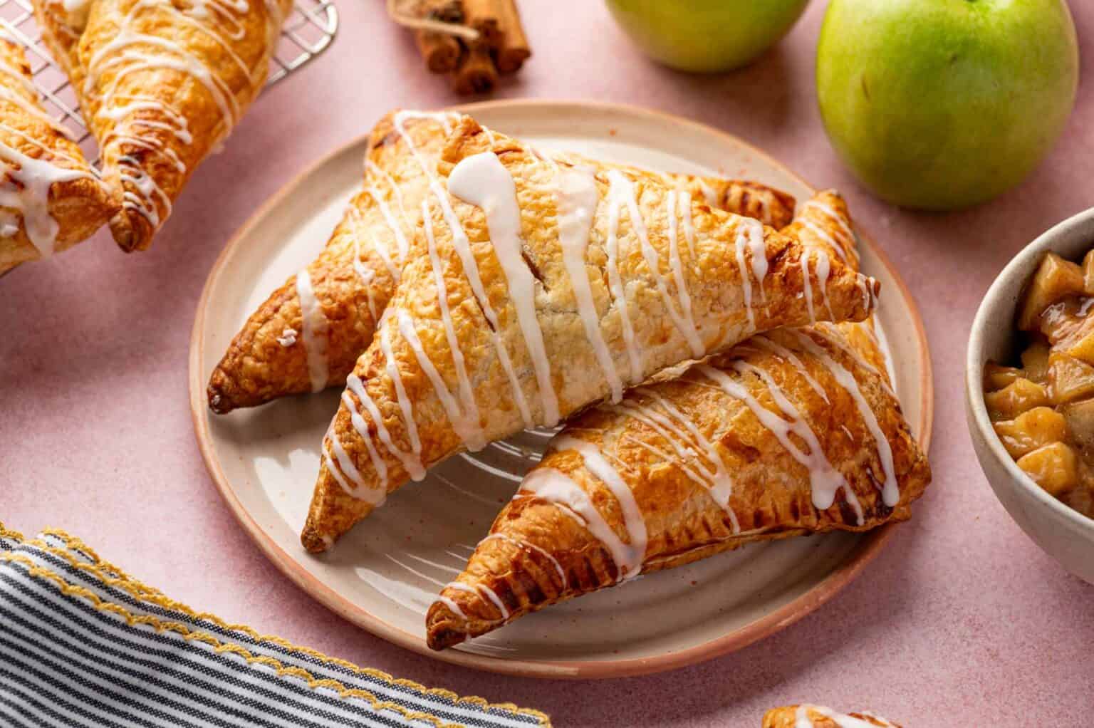 Three golden-brown, triangular apple turnovers with white icing are stacked on a plate, surrounded by green apples and a bowl of apple filling—an easy dessert that’s perfect for kids or as a sweet addition to your favorite autumn desserts.