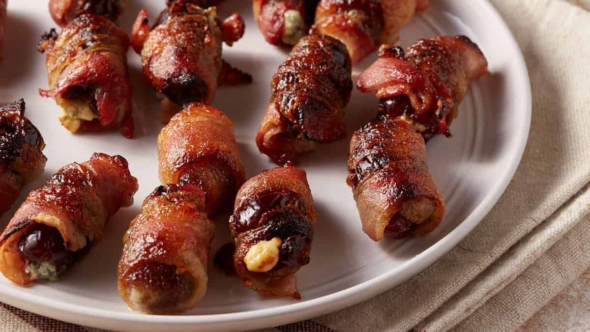 A white plate holds several bacon-wrapped dates, some with visible stuffing, arranged in a loose cluster—perfect party appetizers for any gathering.