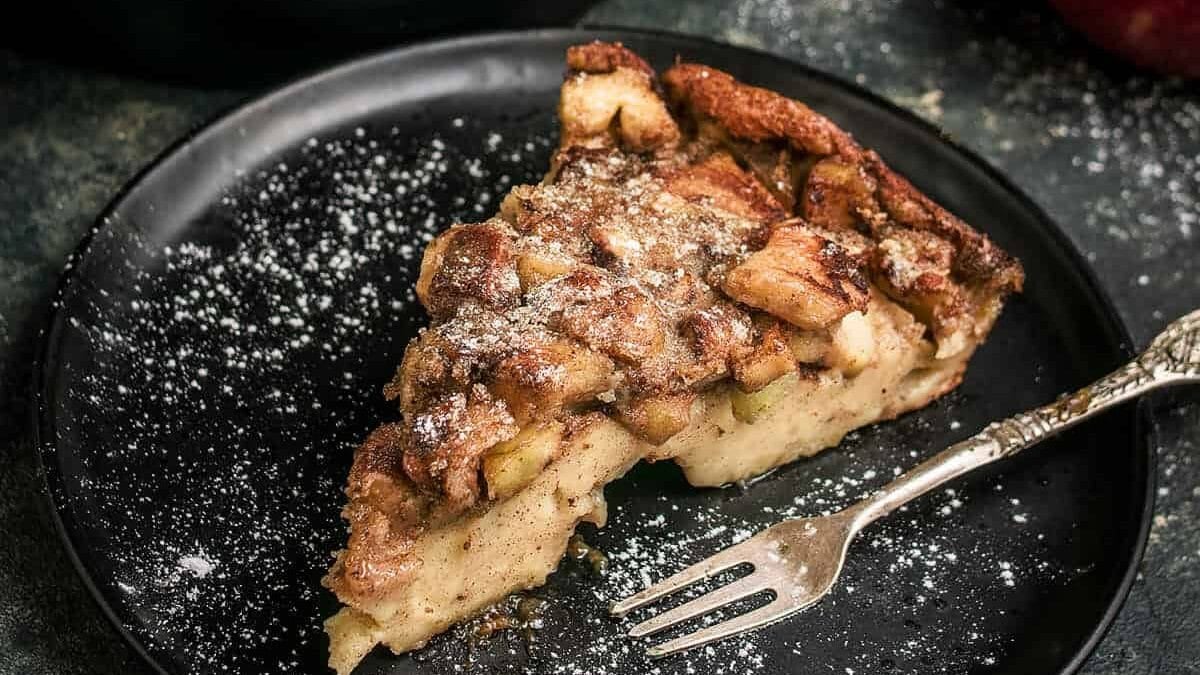 A slice of apple bread pudding on a black plate, dusted with powdered sugar, with a fork resting beside it—a cozy treat inspired by German Favorites and perfect for your Oktoberfest celebration.