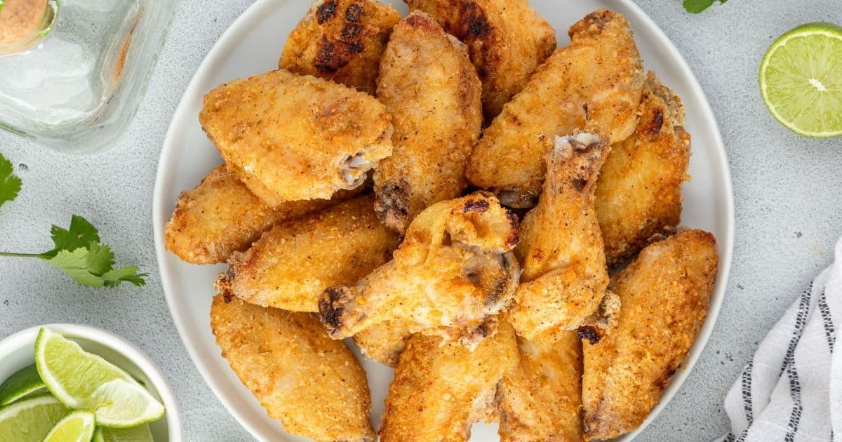 A white plate filled with crispy, seasoned chicken wings sits on a light surface, surrounded by lime wedges and fresh cilantro—perfect Game Day Food.