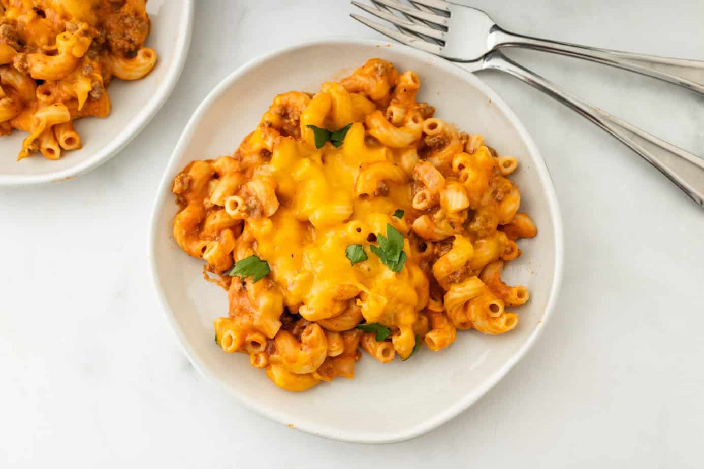 Plate of macaroni and cheese with ground beef, topped with melted cheddar and a few parsley leaves, next to three metal forks on a white surface—perfect for easy family dinners or anyone looking for kid-friendly meals everyone will love.