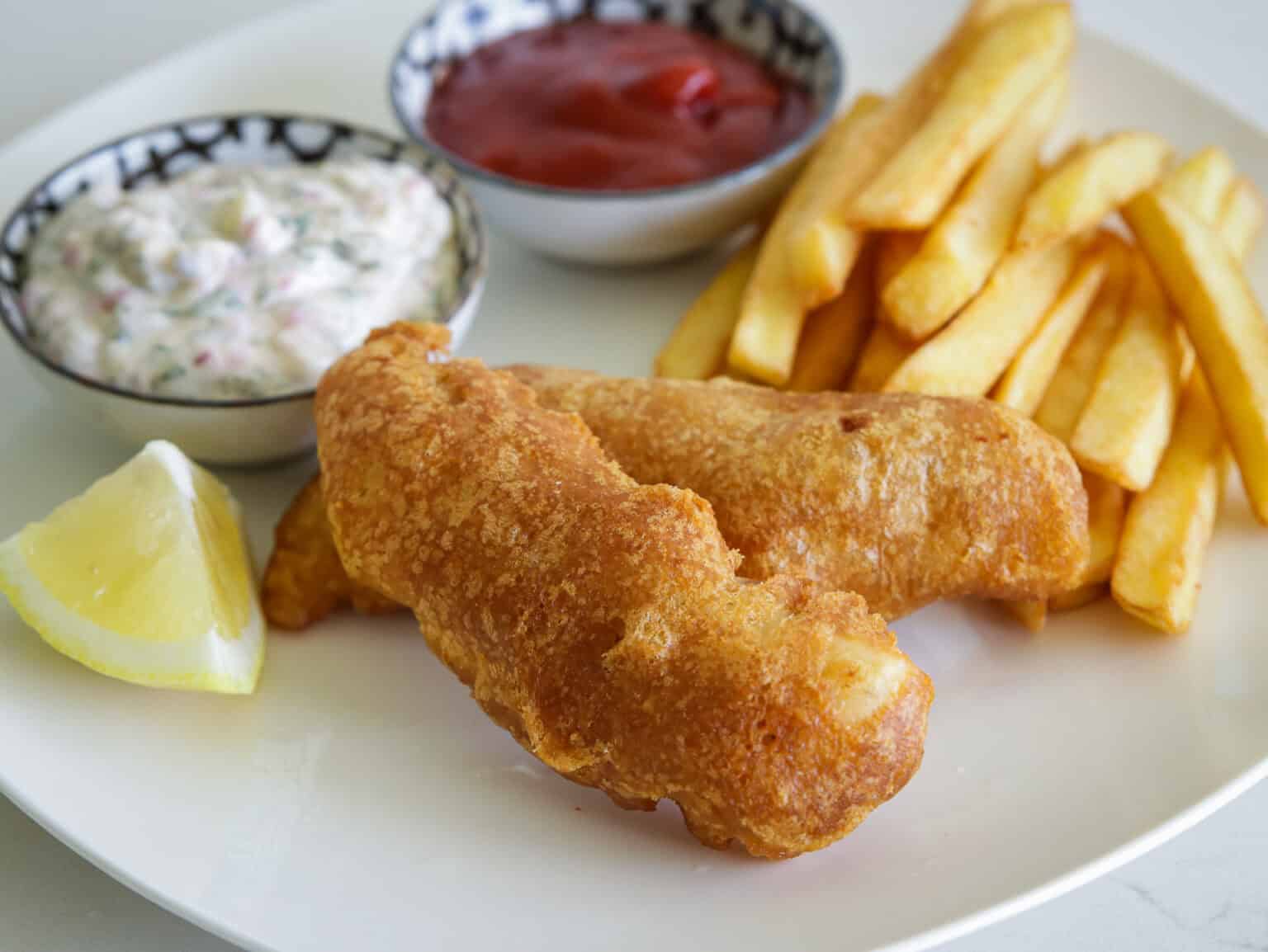 Two pieces of battered fish, French fries, a lemon wedge, tartar sauce, and ketchup served on a white plate—this classic meal stands out among the best fish dishes and is perfect for anyone looking for easy fish recipes to satisfy their cravings.