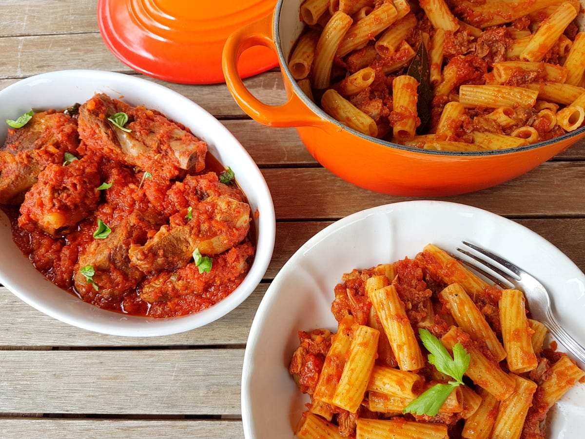 A pot and two bowls of rigatoni pasta with tomato sauce and meat pieces, perfect for Sunday nights and Cozy braises, garnished with fresh herbs on a wooden table.