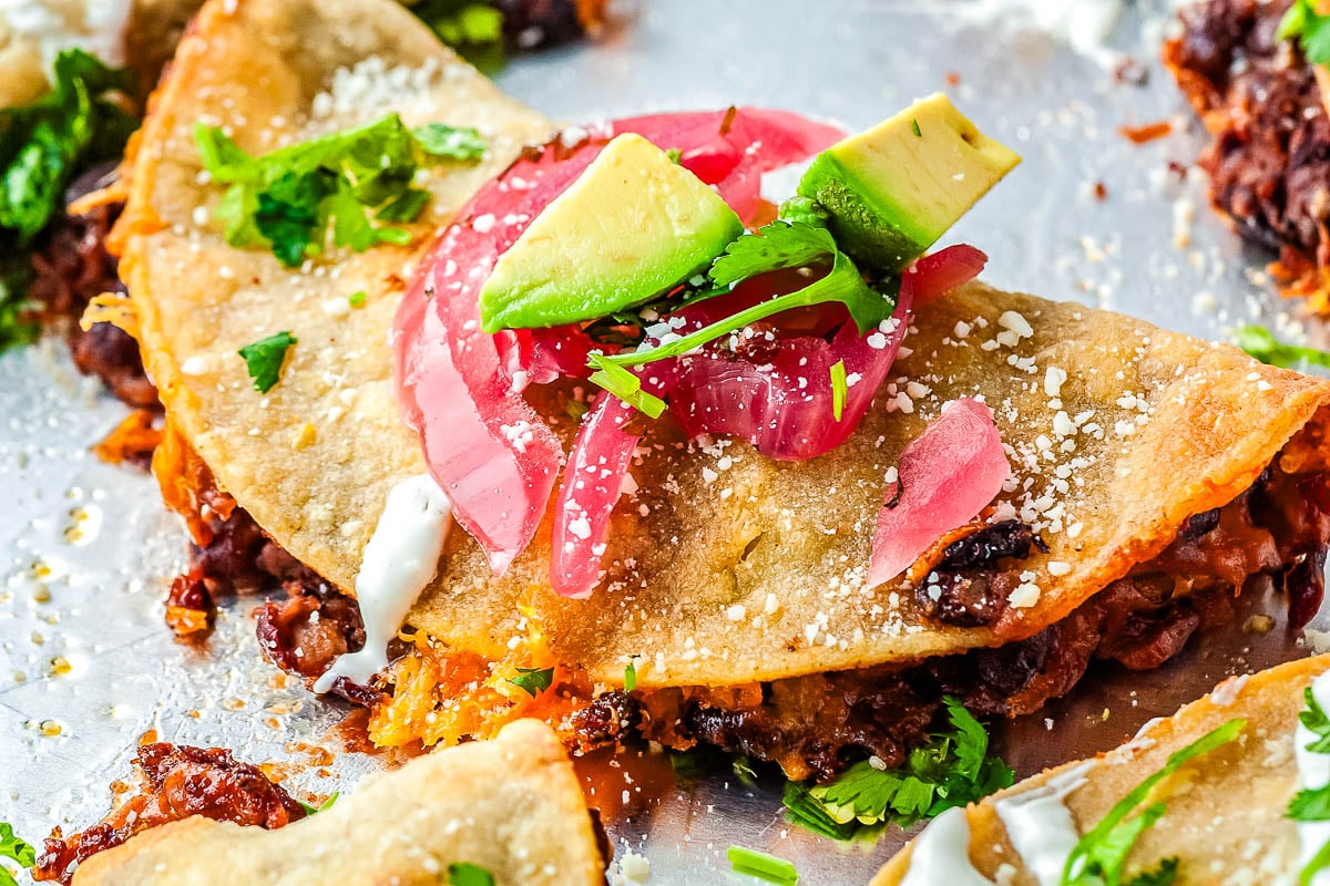 A crispy taco filled with beans and cheese, topped with avocado slices, pickled onions, cilantro, white sauce, and crumbled cheese, served on a metal tray—a perfect Tex Mex dinner inspired by classic Tex Mex recipes.