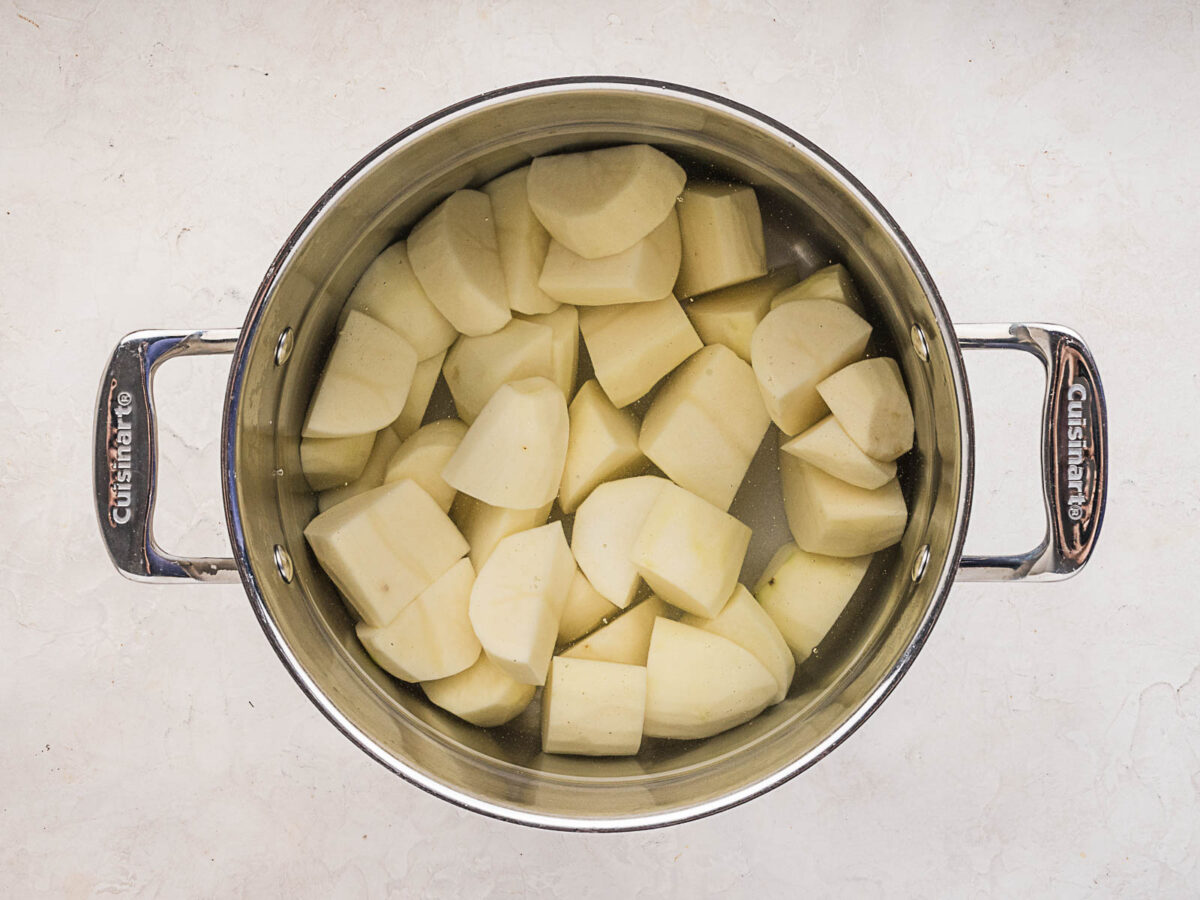 A stainless steel pot filled with peeled, chopped potatoes submerged in water, viewed from above on a light countertop.