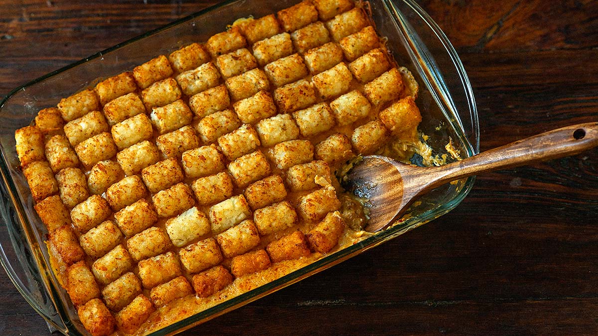 A glass baking dish filled with tater tot casserole—an easy meal perfect for dinner recipes—with a wooden spoon resting inside and some of the casserole scooped out.