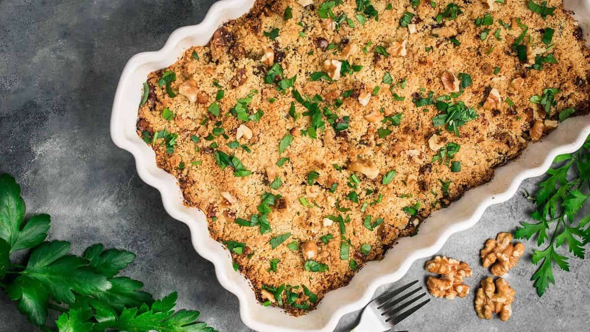 Rectangular baking dish with a golden-brown breadcrumb and herb topping, garnished with chopped parsley and walnuts, on a gray surface with fresh parsley and walnuts nearby—a delicious addition to your collection of unique casserole recipes. Save for later to inspire your next weeknight dinner!.