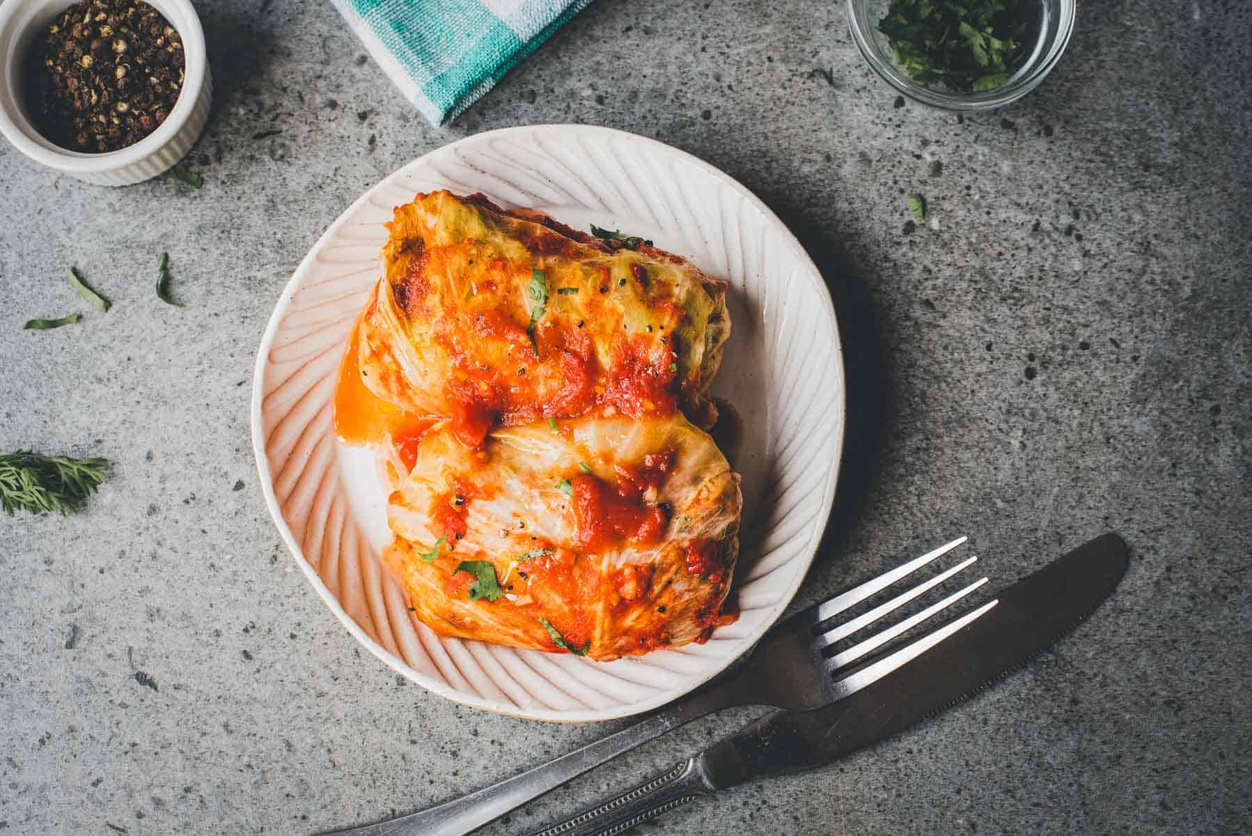 A plate with two pieces of baked stuffed cabbage rolls topped with tomato sauce, reminiscent of classic casserole recipes, with a fork and knife beside the plate on a gray countertop.