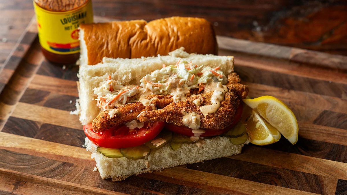 Fried catfish po’ boy sandwich with tomatoes, pickles, coleslaw, remoulade sauce, and lemon wedges on a wooden board, with a Louisiana hot sauce bottle in the background—a true taste of Southern food perfect for any Southern Supers gathering.