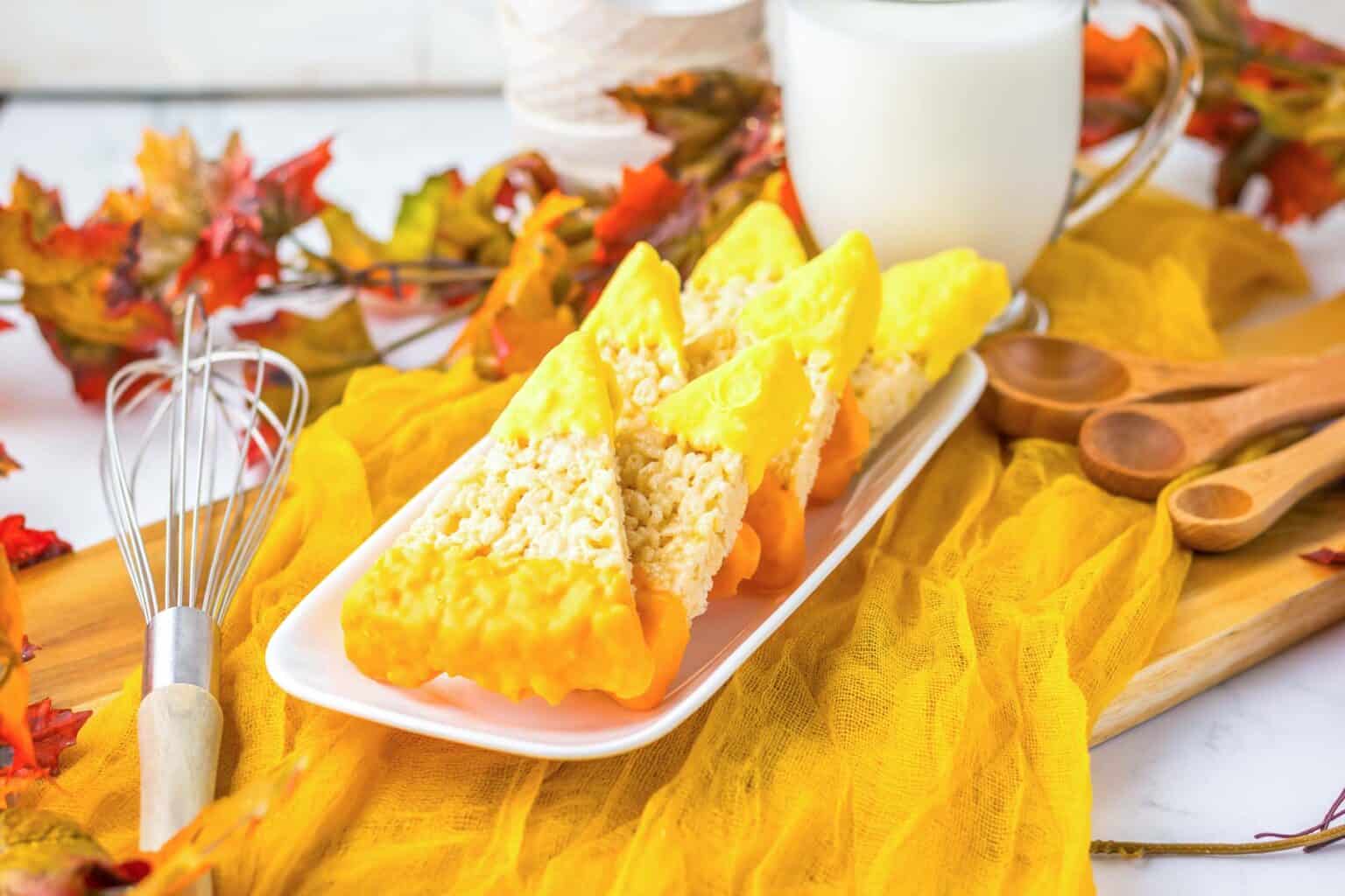 Three triangular rice crispy treats dipped in yellow and orange icing sit on a white plate, accompanied by a glass of milk, autumn leaves, and kitchen utensils. These easy desserts are perfect for kids and make delightful autumn desserts the whole family will enjoy.