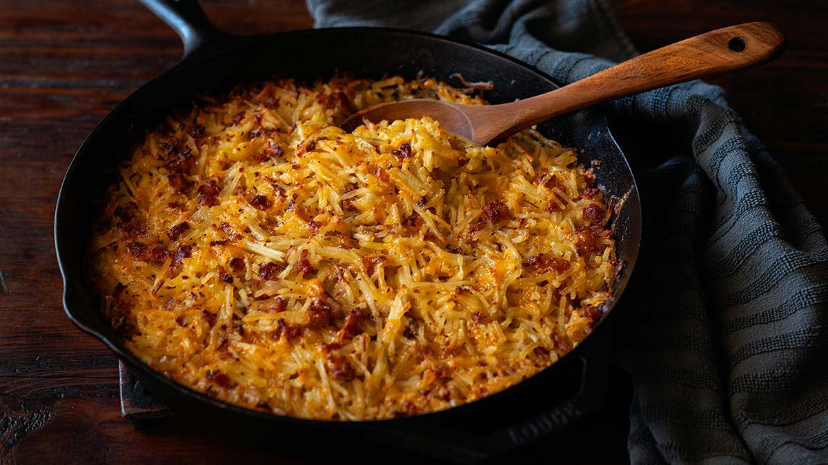 A cast iron skillet filled with baked hash brown casserole topped with melted cheese and bits of bacon, with a wooden spoon resting inside—one of the best potluck recipes to feed a crowd, and guaranteed to make hosting stress free.