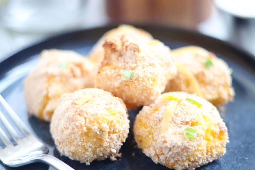 Six breaded, round cheese balls garnished with green herbs and cheese are arranged on a black plate next to a fork, making them a delicious addition to your collection of potato side dishes for a cozy fall dinner.