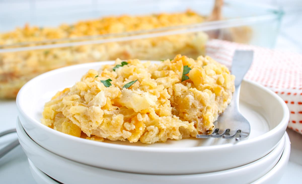A serving of cheesy hash brown casserole on a white plate with a fork, garnished with parsley, with a casserole dish in the background—perfect for anyone searching for delicious and easy casseroles to add to their collection of dinner recipes.