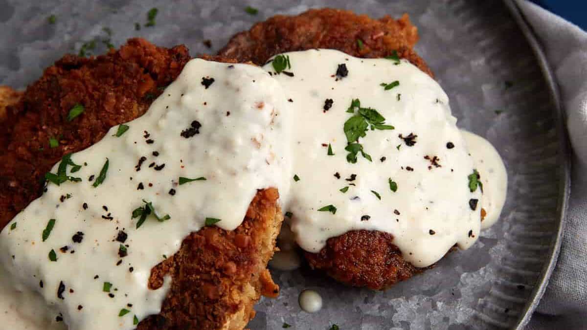 Two pieces of breaded fried steak covered in white pepper gravy and garnished with chopped parsley on a gray plate—classic comfort food that brings the warmth of home cooking to your table, perfect for hearty Sunday suppers.