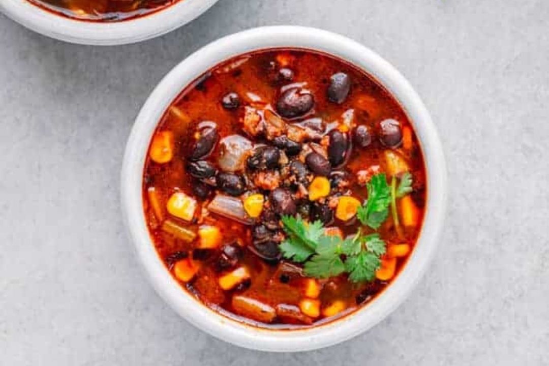 A bowl of chili with black beans, corn, onions, and herbs in a red sauce, garnished with cilantro, on a gray surface—perfect for those who love comforting soups and crave delicious bean soup recipes.