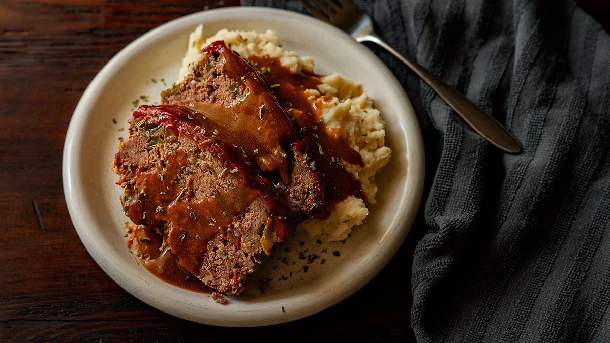 A plate of meatloaf topped with brown gravy and herbs, served over mashed potatoes, with a fork on the side—this is one of those classic recipes everyone should learn for true comfort food at home.