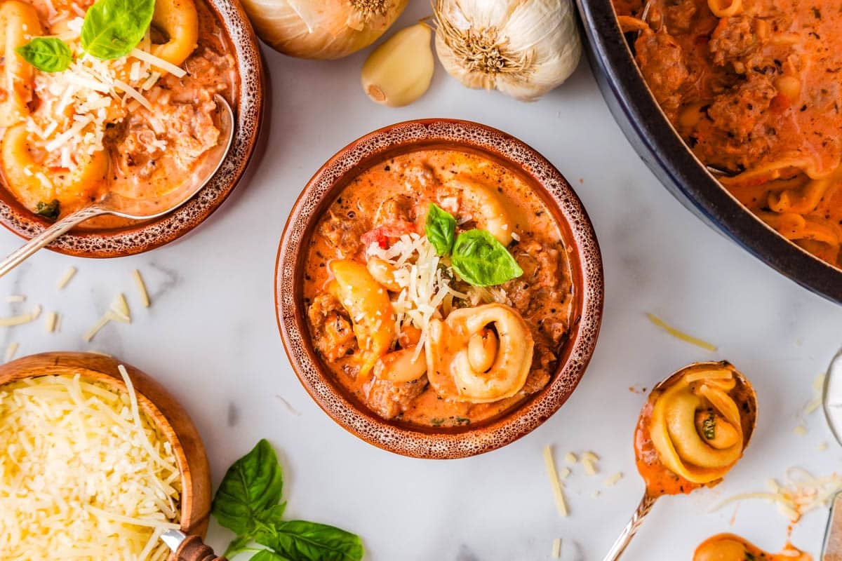 Bowl of creamy tortellini soup with sausage, topped with grated cheese and basil, surrounded by ingredients like cheese, basil, onion, and garlic on a white surface—perfect for anyone looking for cozy fall recipes or comforting creamy soup recipes.