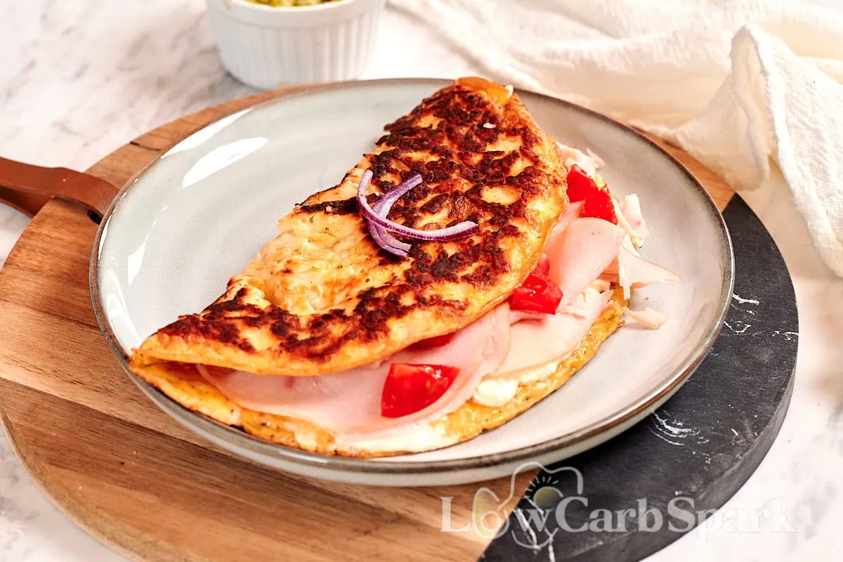 A folded omelet on a plate, filled with sliced turkey, tomato pieces, and red onion, set on a round wooden board with a small bowl in the background—perfect for those who love savory breakfast recipes.