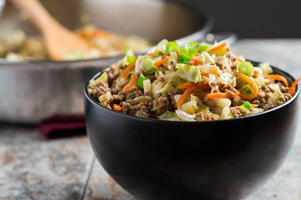 A black bowl filled with a savory stir-fry featuring ground meat, shredded cabbage, carrots, and chopped green onions—a delicious example of easy cabbage dishes. In the background, a skillet with more food and a wooden spoon completes this mouthwatering cabbage recipe scene.