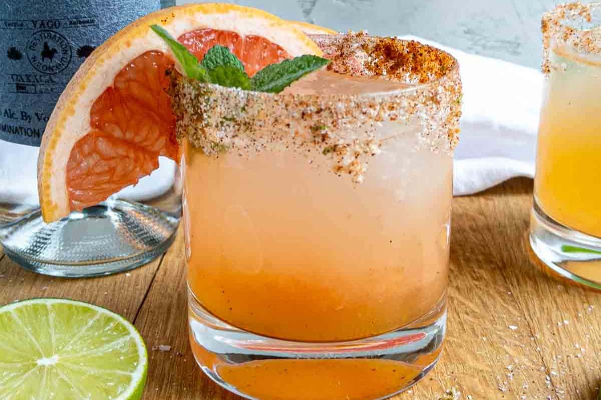 A glass of grapefruit cocktail with a chili-salt rim, garnished with a grapefruit slice and mint, sits on a wooden surface next to a lime half—perfect for those seeking refreshing brunch cocktails or looking to try new cocktail recipes.