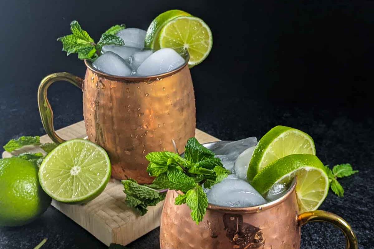 Two copper mugs filled with ice, lime slices, and mint leaves—perfect for brunch cocktails—are set on a dark surface with additional limes and mint nearby.