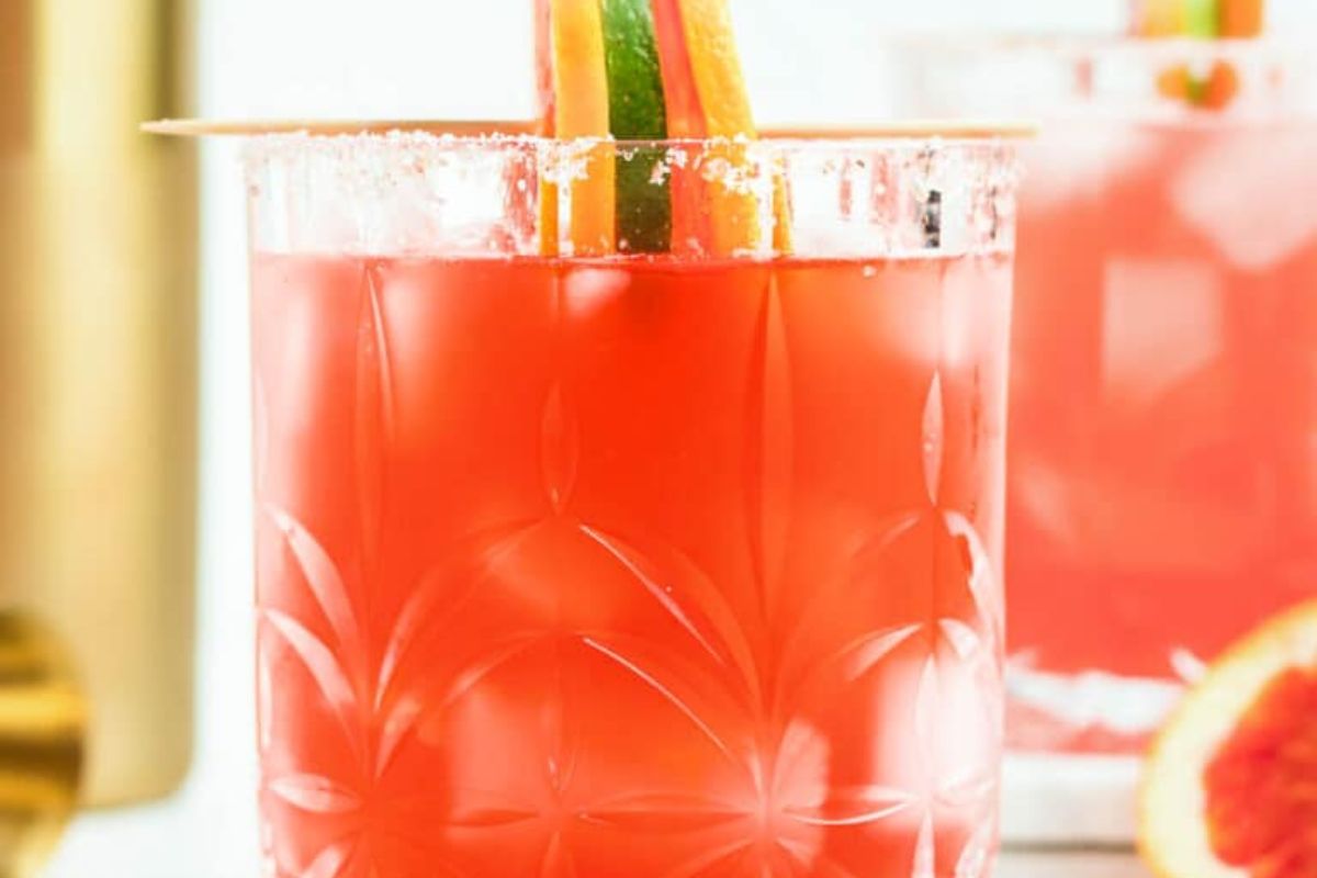 A glass of red cocktail with a salted rim, garnished with slices of lime and orange, sits on a table—an inviting choice for anyone seeking new brunch cocktails to enjoy with friends.
