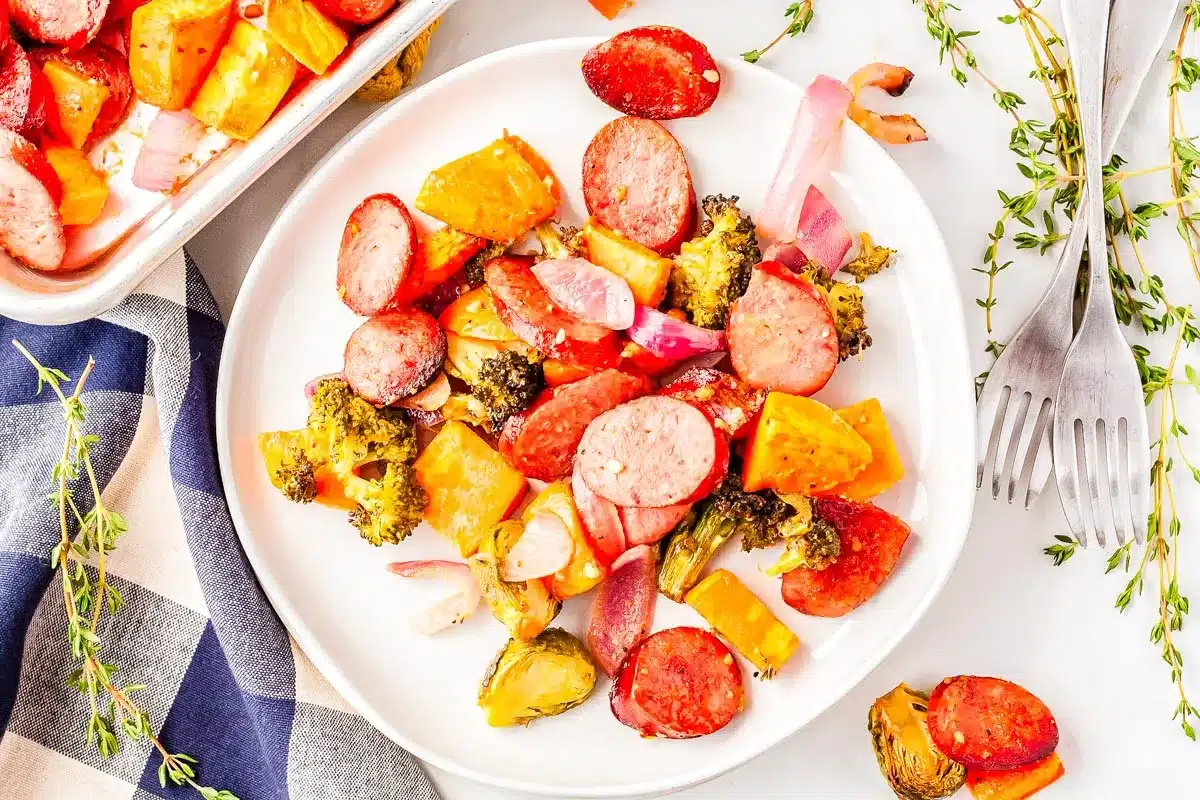 A white plate with sliced sausage, broccoli, sweet potatoes, red onions, and brussels sprouts. Perfect for those who love trying new savory recipes, this colorful dish is arranged next to two forks and fresh thyme on a white surface.