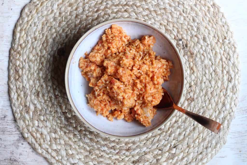 A bowl of orange-colored rice with visible grains and chunks, placed on a round woven placemat with a spoon—this is one of those flavorful rice dishes that perfectly balances taste and presentation.