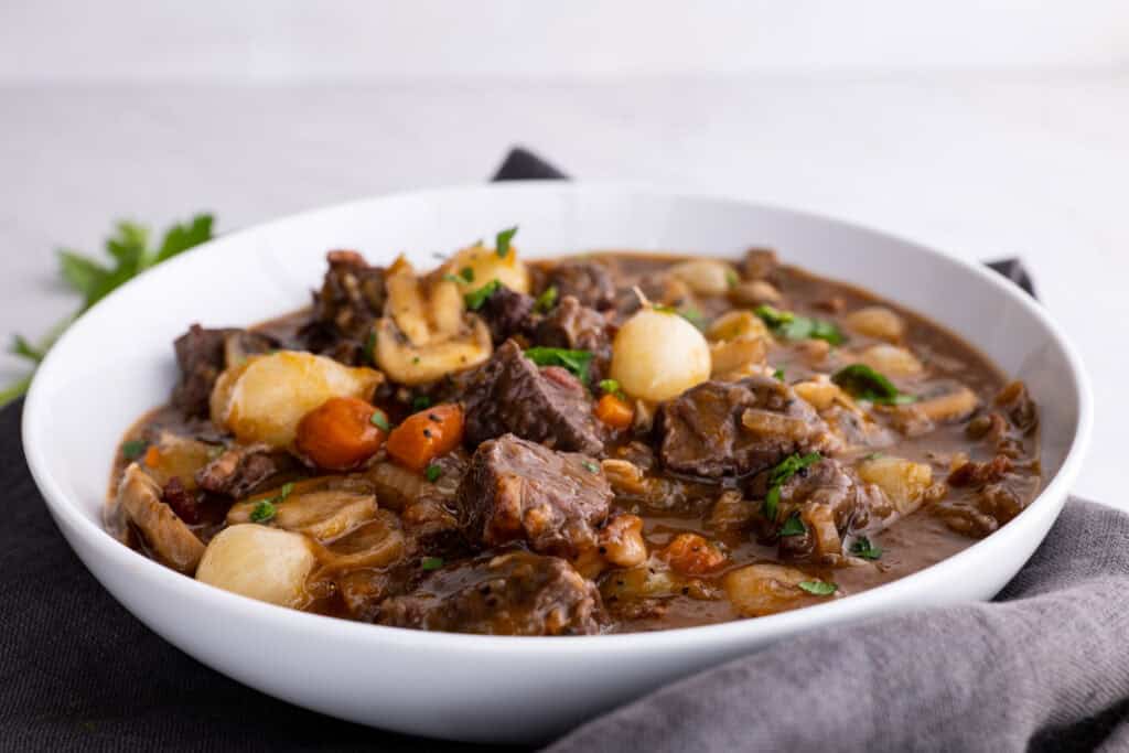 A white bowl filled with beef stew, featuring chunks of meat, carrots, mushrooms, pearl onions, and a thick brown sauce—perfect for cozy braises on Sunday nights.
