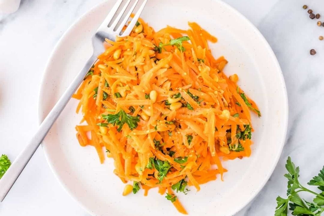 Shredded carrot salad with parsley and seeds is served on a white plate with a fork, making it one of the best quick carrot sides for any meal.