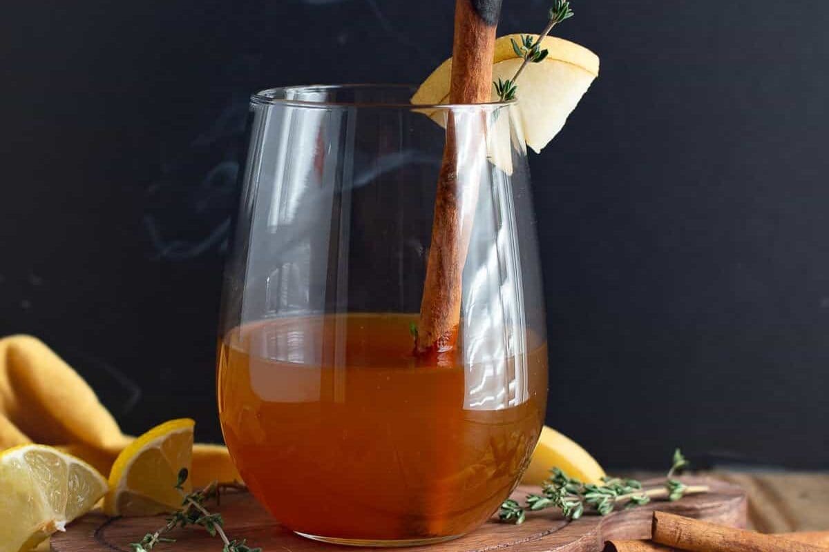A glass of spiced apple cider garnished with a cinnamon stick, apple slice, and thyme sprig, with lemon wedges and herbs on a wooden surface—an inviting twist on classic brunch cocktails that promises to elevate your collection of cocktail recipes.