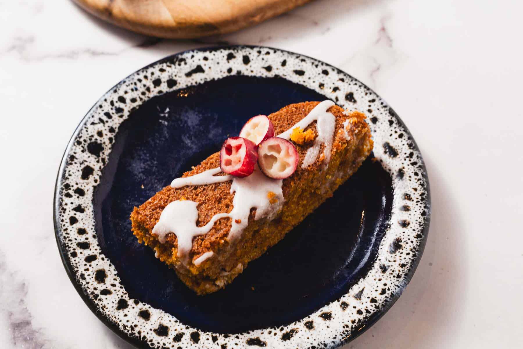 A rectangular slice of gluten free cake with white icing and halved cranberries on top, served on a dark plate with a speckled rim—perfect for fans of gluten free desserts.