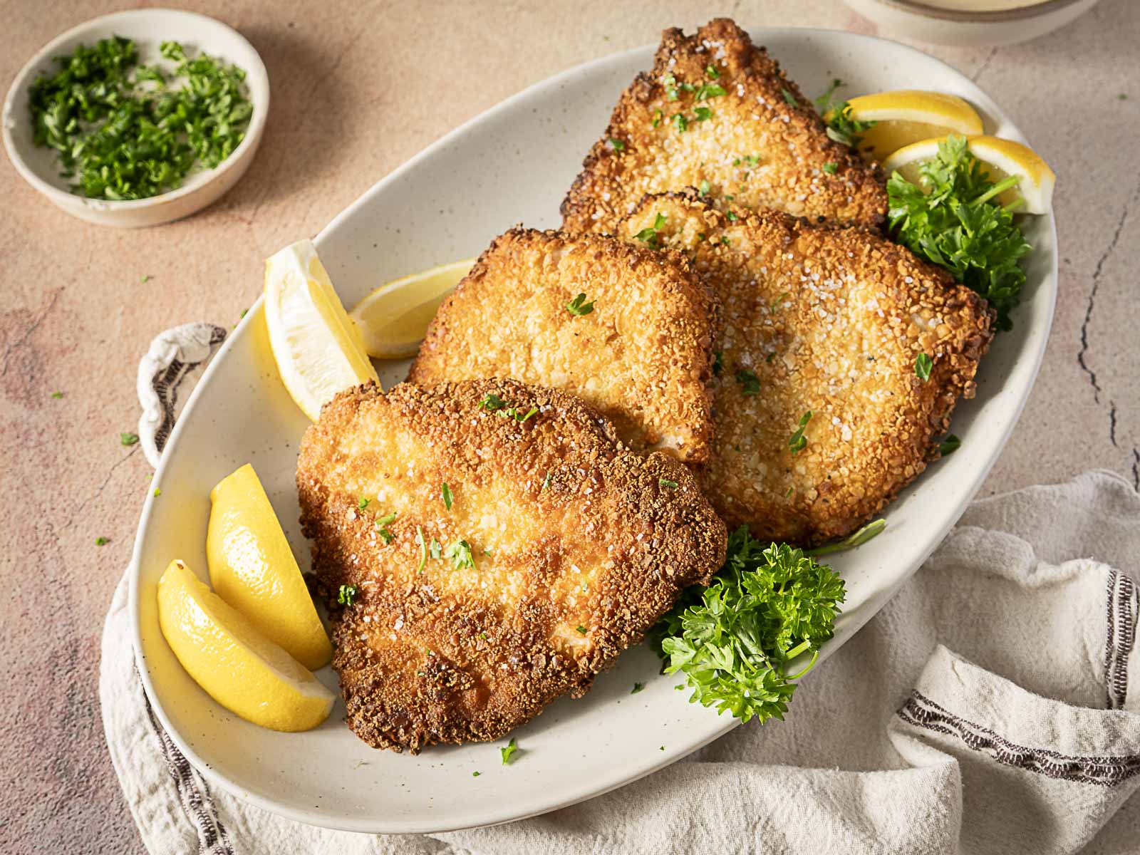 A white plate with three breaded, fried cutlets garnished with chopped herbs, parsley sprigs, and lemon wedges—perfect for those seeking delicious Gluten Free recipes to enjoy among their Fall Favorites.