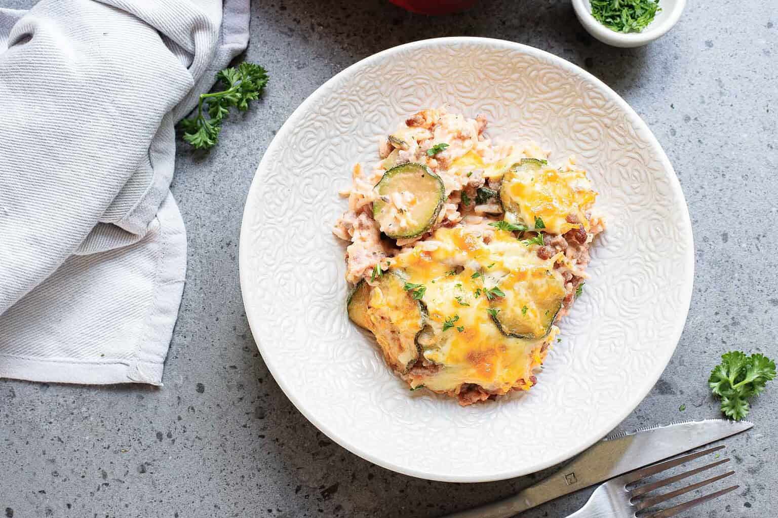 A white plate with a serving of cheesy casserole containing rice and zucchini, placed on a gray countertop with a fork and napkin nearby—a perfect choice for those seeking easy casseroles when looking for quick meals or comforting dinner recipes.