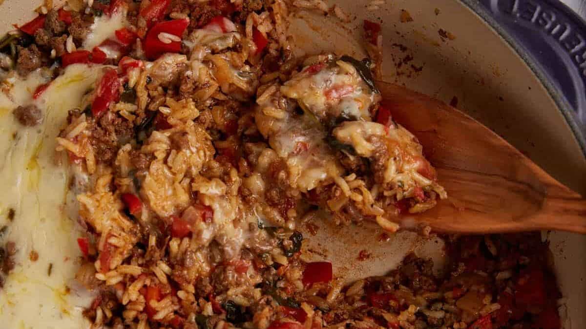 A close-up of a baked rice and ground meat casserole with melted cheese and red peppers, being stirred with a wooden spoon in a pot—a perfect example of flavorful rice dishes for anyone seeking simple rice recipes.