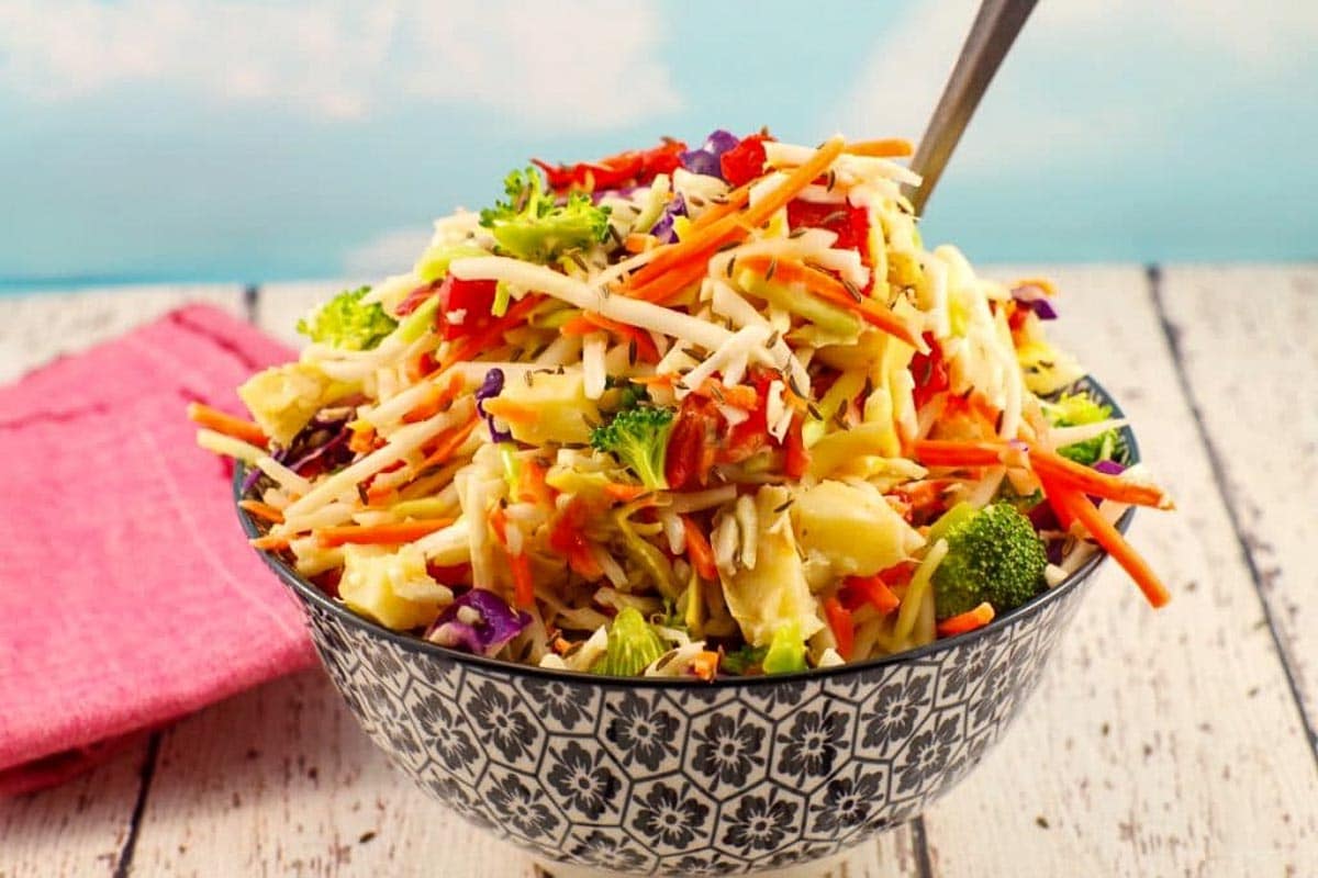 A patterned bowl filled with coleslaw made of shredded carrots, cabbage, broccoli, and red bell peppers—perfect as a vibrant side for roasted pepper dishes—sits on a wooden table with a pink napkin.