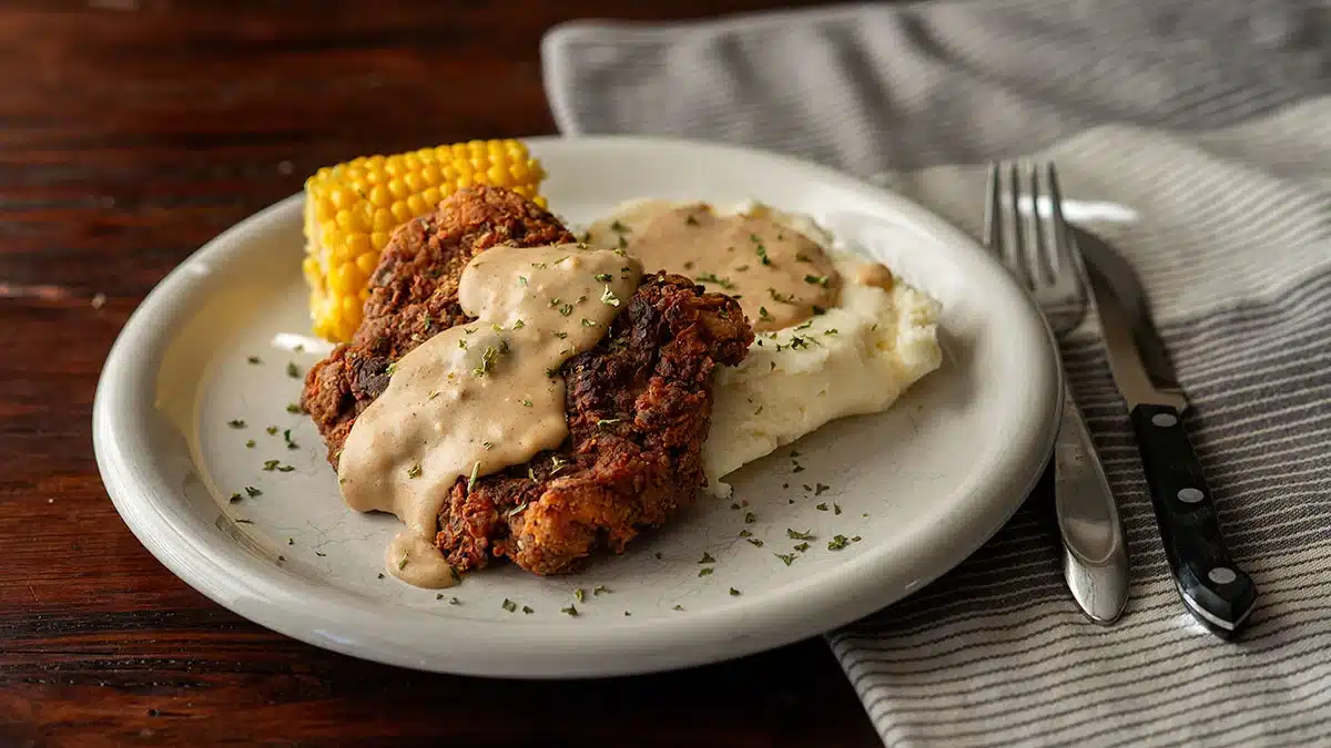 Plate with chicken fried steak topped with gravy, mashed potatoes, and a piece of corn on the cob, set on a wooden table next to a striped napkin with a fork and knife—a delicious example of classic recipes enjoyed for generations.