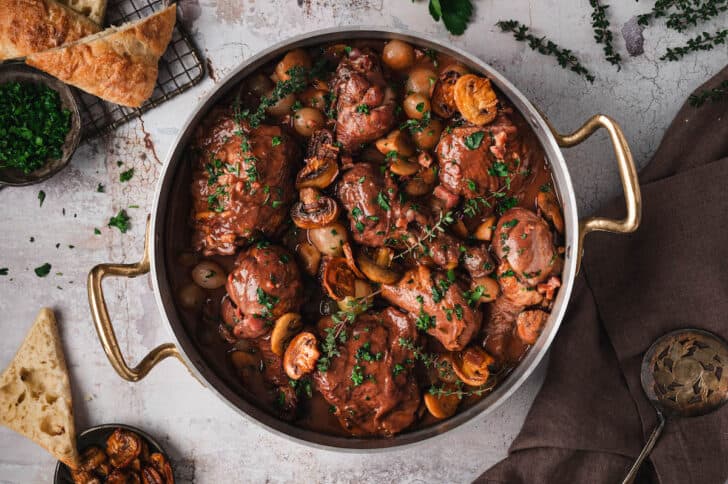A pot of coq au vin with chicken, mushrooms, onions, and herbs sits on a table—perfect for cozy braises on Sunday nights, surrounded by bread slices and chopped parsley.