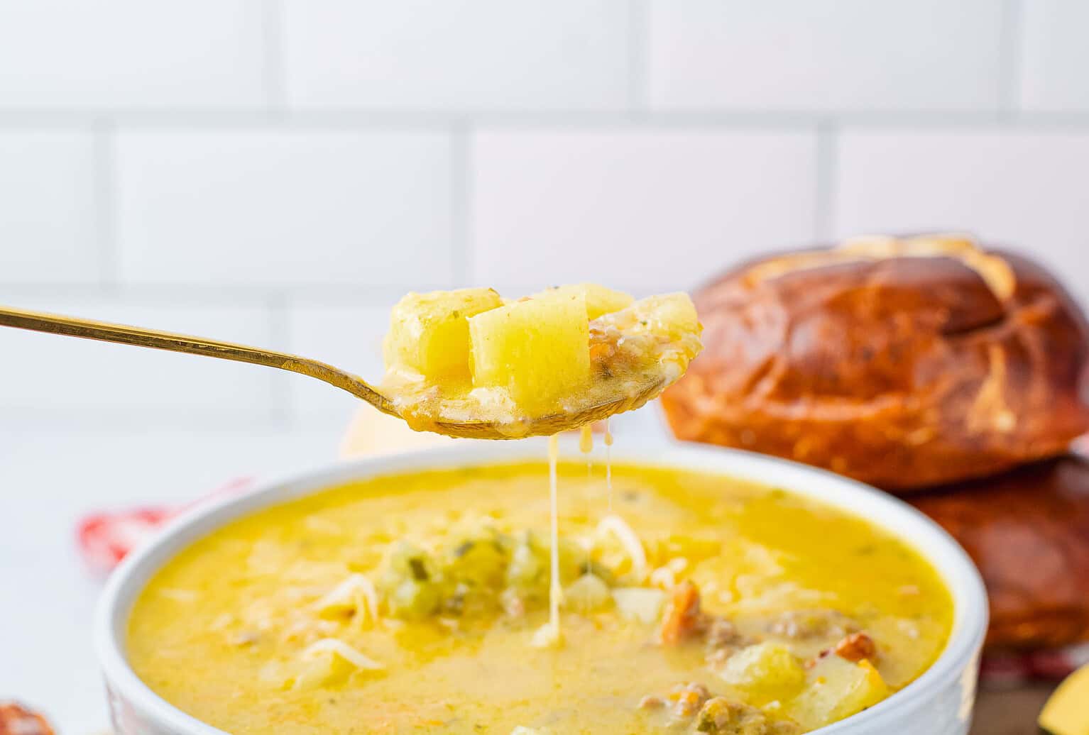 A spoonful of cheesy potato soup is held above a bowl, with bread rolls in the background on a white tiled surface—perfect for cool nights and those seeking cozy soup recipes this fall.