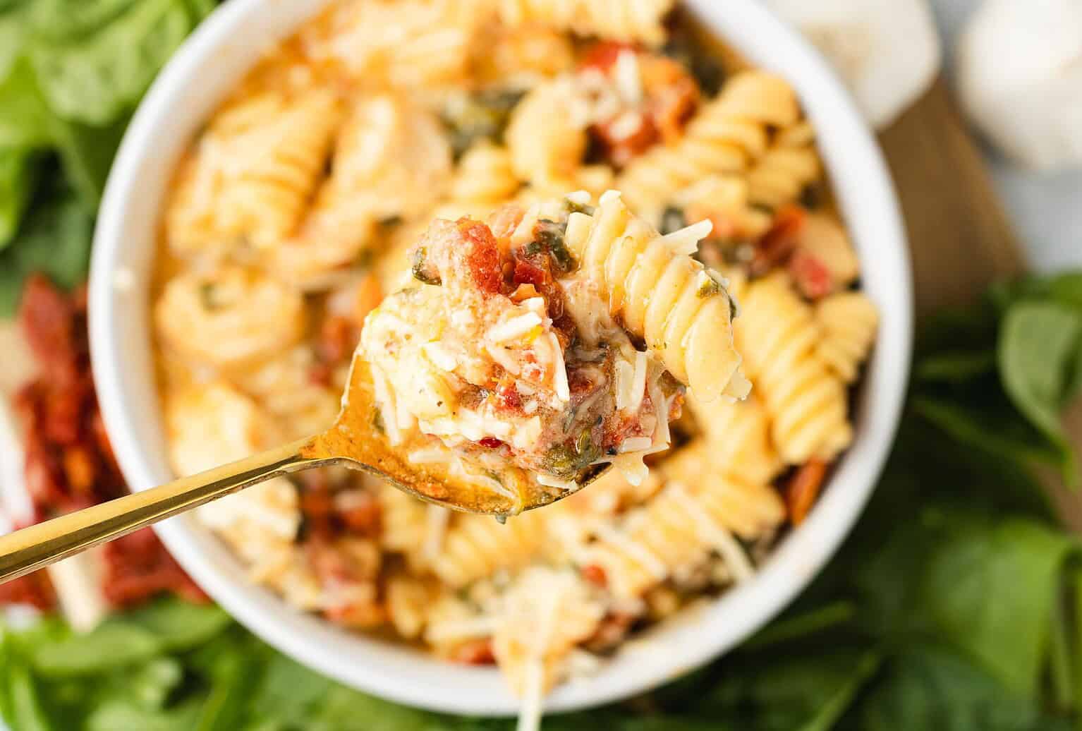 A close-up of a fork holding a bite of rotini pasta with cheese, tomatoes, and herbs over a bowl of the same pasta. Fresh greens surround the bowl, setting the perfect scene for fall and cool nights when cozy soup recipes and comforting dishes are in high demand.