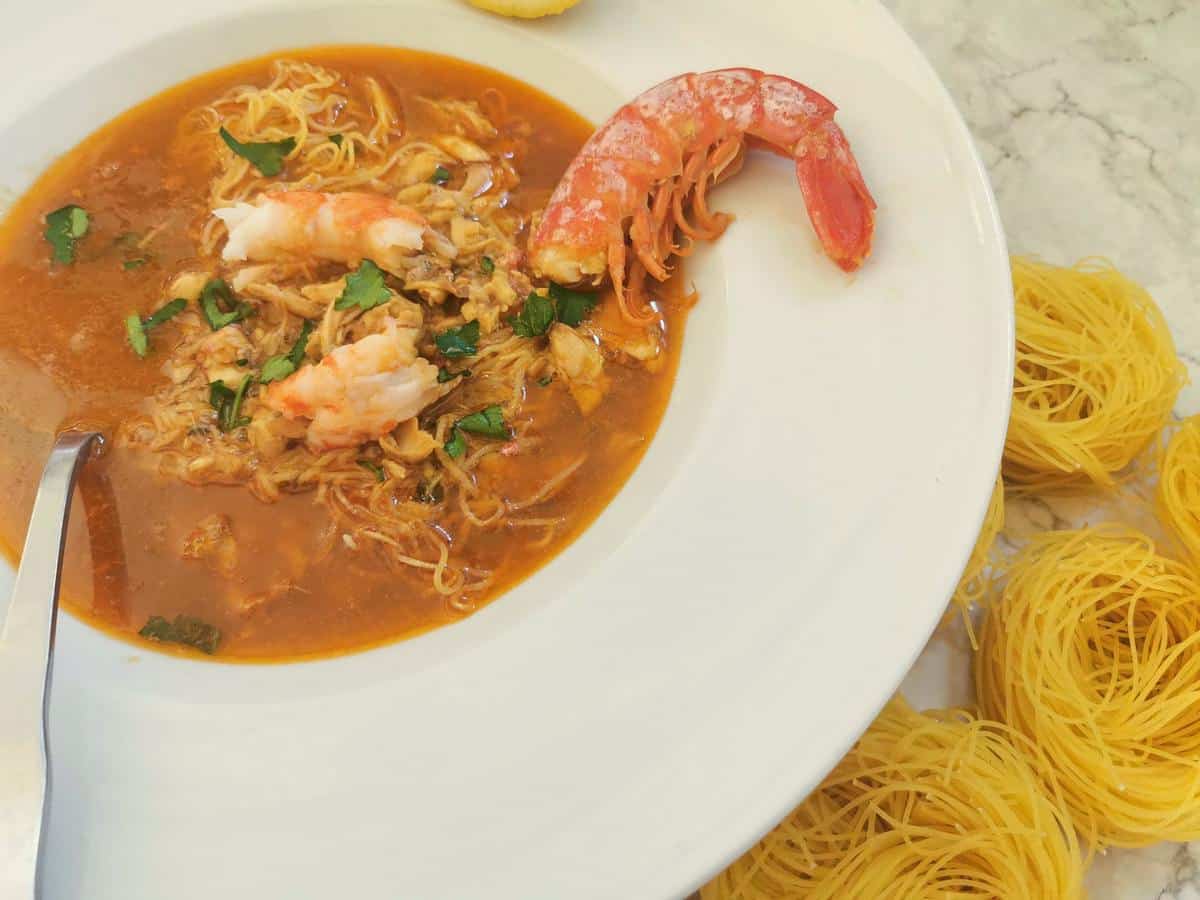 Bowl of shrimp noodle soup in red broth on a white plate with a spoon, garnished with herbs; uncooked yellow noodles beside the plate—a delicious addition to your collection of easy fish dishes and seafood meals.