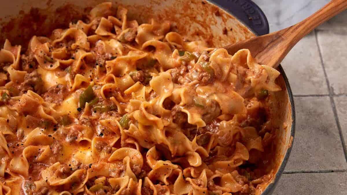 A wooden spoon scoops cheesy ground beef and noodle casserole from a large pot on a tiled surface, making this one of the ultimate easy casseroles for your weeknight dinner recipes collection.