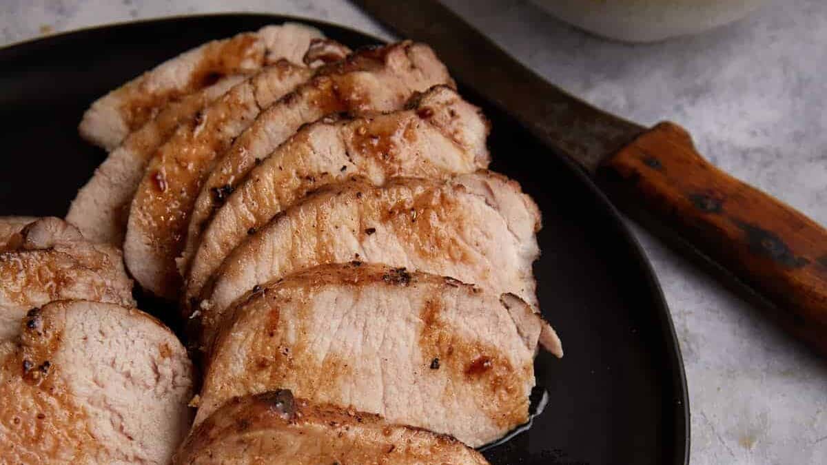 Sliced cooked pork loin arranged on a black plate, with a knife featuring a wooden handle beside it—perfect for savoring with pancakes or drizzling with maple syrup.