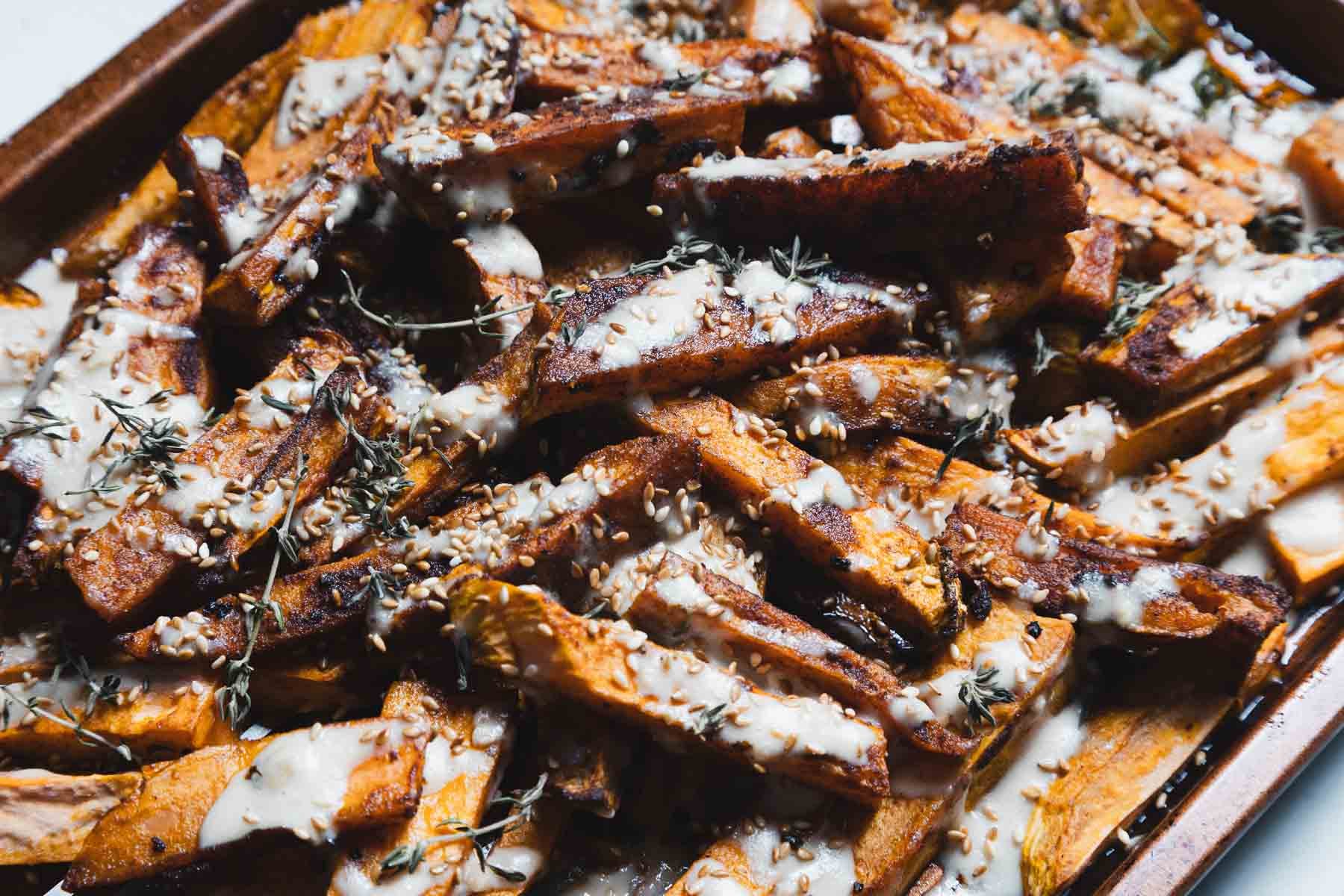 Baked sweet potato fries on a tray, topped with a creamy white sauce, sesame seeds, and fresh herbs—an irresistible addition to your favorite sweet potato recipes.