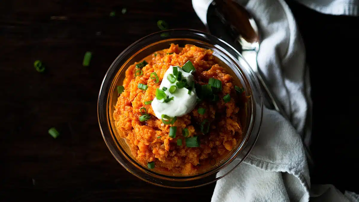 A bowl of mashed sweet potatoes, a delicious alternative sweet potato dish, topped with sour cream and green onions beside a spoon and white napkin on a dark surface.