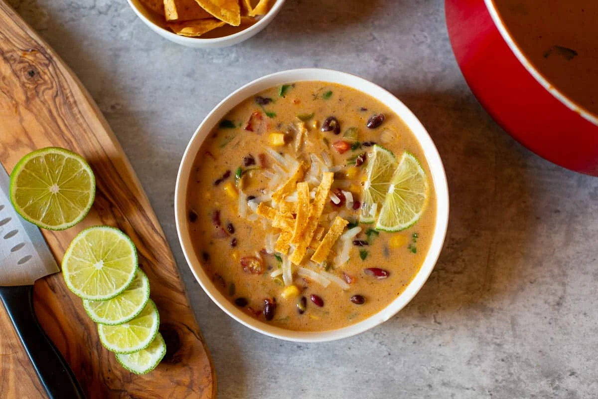 A bowl of creamy soup with beans, corn, tortilla strips, and a lime slice—perfect for fans of comforting soups—sits next to sliced limes and a pot on a gray surface.